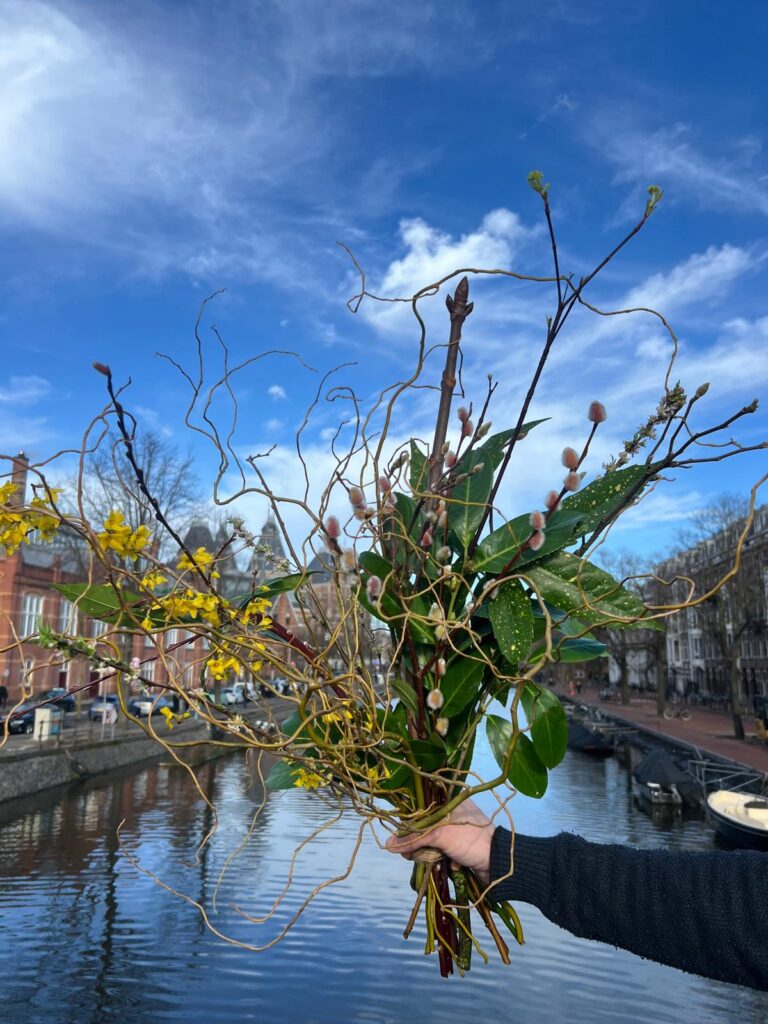 Op de achtergrond het rijksmuseum, boven het water houd de bloemist een boeket vast van bloeiend hout.